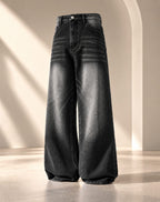Wide-leg black denim jeans with a faded gray wash and horizontal seam detailing, displayed on a neutral beige studio set with soft architectural shadows.